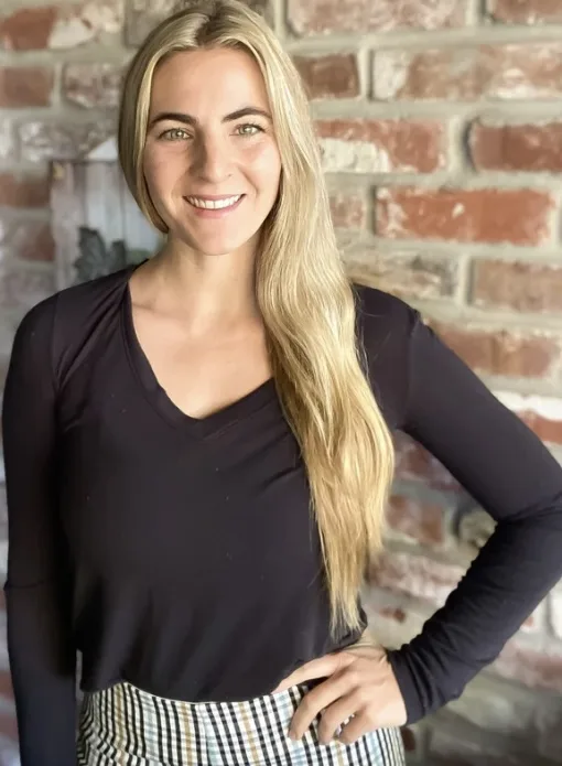 Portrait of Stacey Ring smiling, with long blonde hair, wearing a black top in front of a brick wall